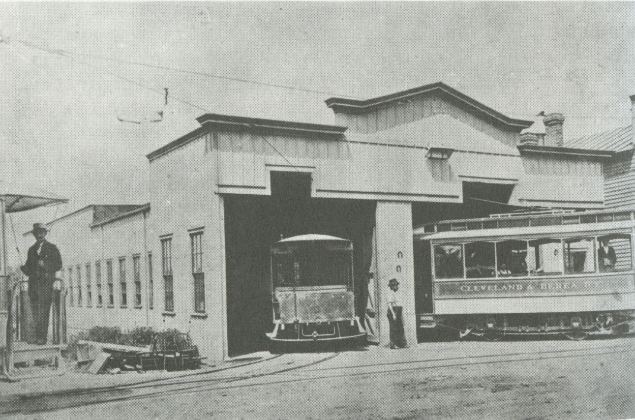 Berea Car Barn Circa the 1890s. One streetcar is parked while the other is in motion. A few workers pose for the photography. Text: "Cleveland & Berea Railway"