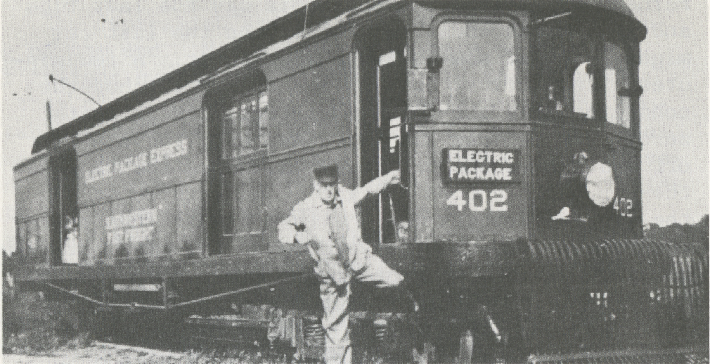 Streetcar number 402, an Electric Package Agency vehicle. An employee of the interurban poses for the photograph by kneeling on a step and hanging off of a handle on the streetcar.